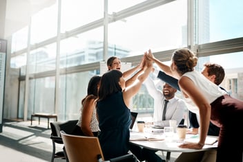Team of six business people high-fiving while sitting in a light and spacious meeting room