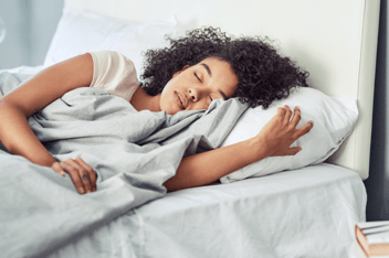 Woman with curly hair sleeping soundly on a comfortable bed with white pillowcase and gray blanket.