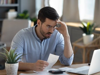 man with his hand on his forehead while holding a receipt and looking at the laptop