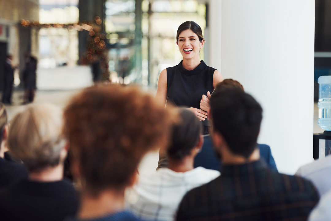 Confident young bookkeeper in black blouse smiling while presenting to a diverse audience during a conference.